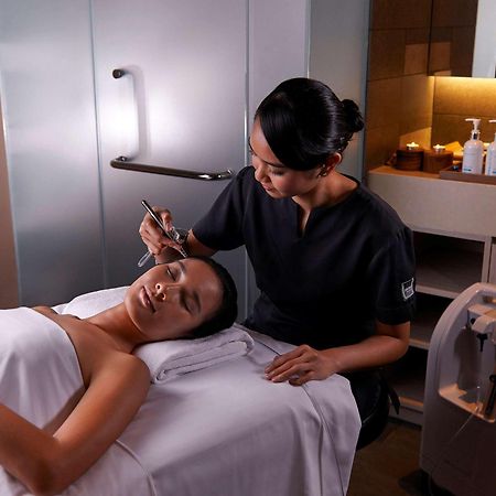Paradox Singapore Merchant Court At Clarke Quay Exterior foto The photo shows a spa or beauty therapy setting where a woman is lying on a treatment table with a towel draped over her. A professional, dressed in a dark uniform, is performing a skincare treatment on her face. Various beauty products are visible o